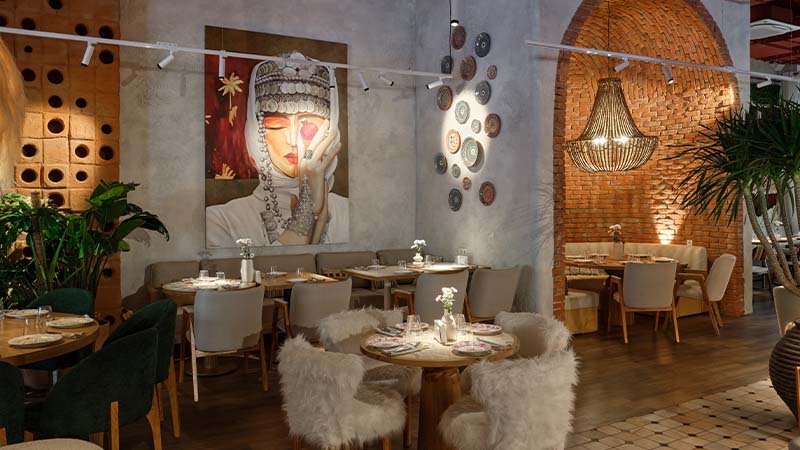 A stylish restaurant interior with wooden tables, plush chairs, a large wall painting of a woman, decorative wall plates, and warm lighting with exposed brick accents.