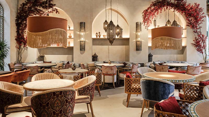 Elegant restaurant interior with round tables, cushioned chairs, hanging fringed lamps, archways, and red foliage decor. Warm lighting creates a cozy, inviting atmosphere.