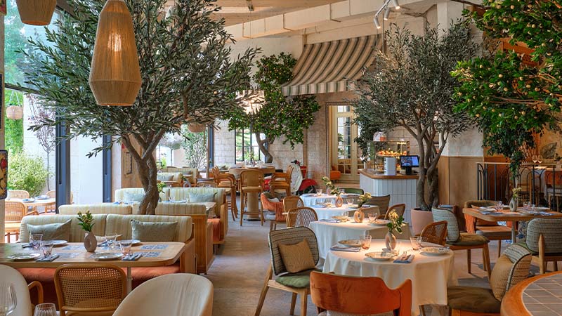 A spacious, well-lit restaurant features indoor trees, wicker chairs, round tables set for dining, and large windows allowing natural light inside.