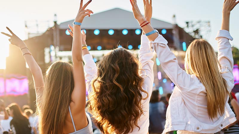 Three people with raised arms enjoy a music festival near a stage.