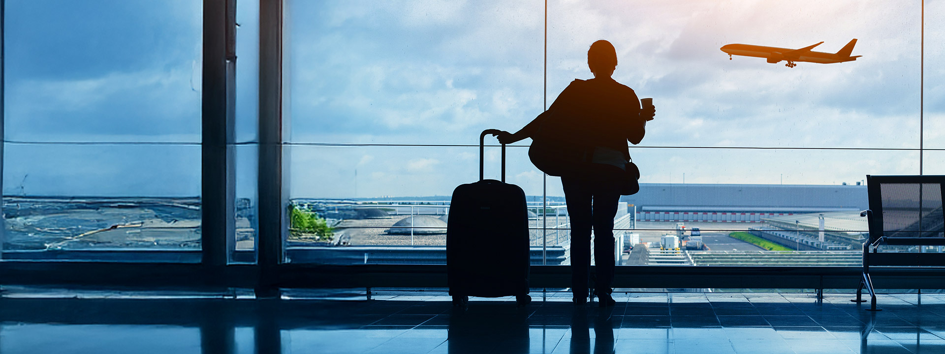 A person stands in an airport terminal with a suitcase, watching an airplane take off through large windows.
