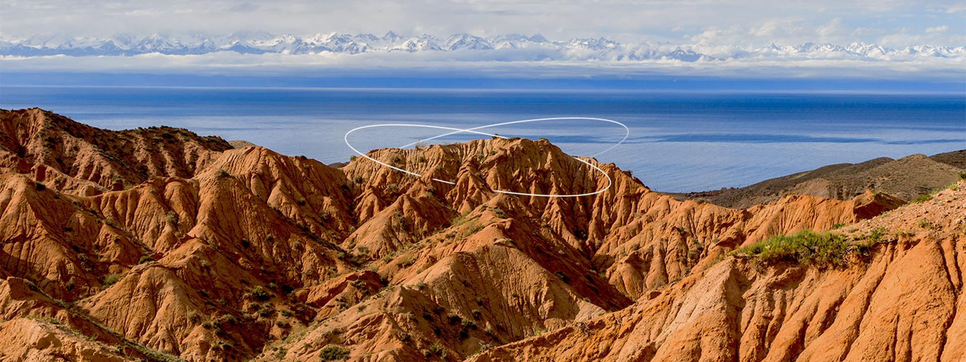 Red rocky hills in the foreground with white lines drawn over a peak, a blue lake in the middle, and snow-capped mountains in the distant background under a cloudy sky.