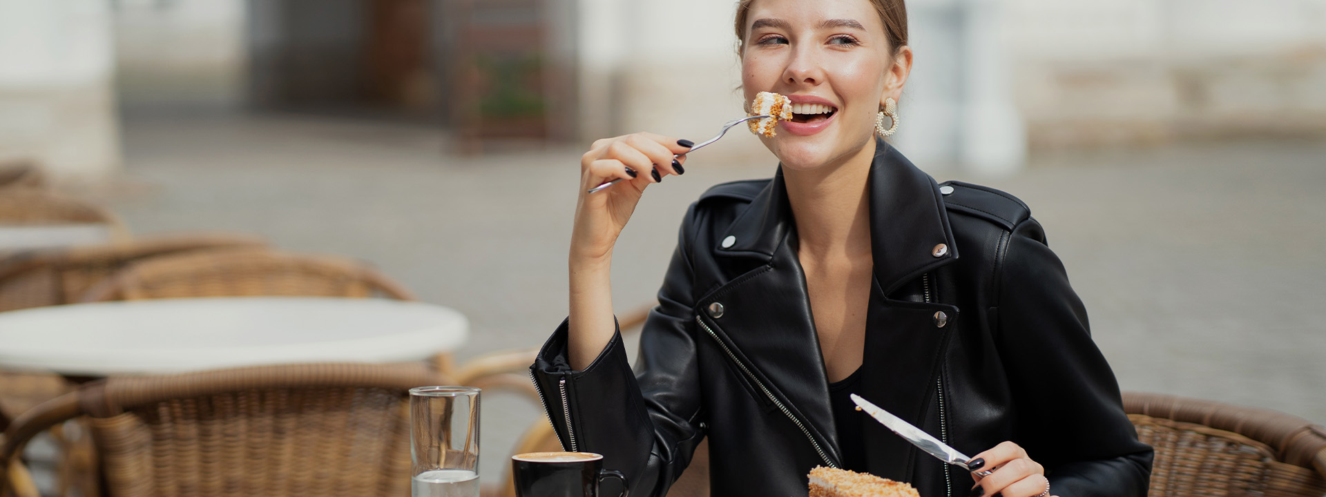 A young woman in a leather jacket eating cake at a cafe table, smiling, with a cup of coffee and a dessert fork in her hands.