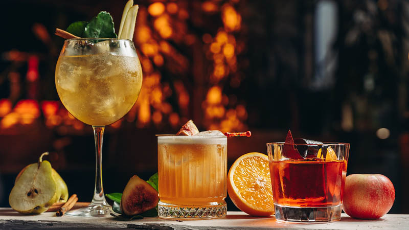 Three cocktails on a bar counter with fresh fruits including pear, fig, orange, and apple in a warm, dimly-lit setting.