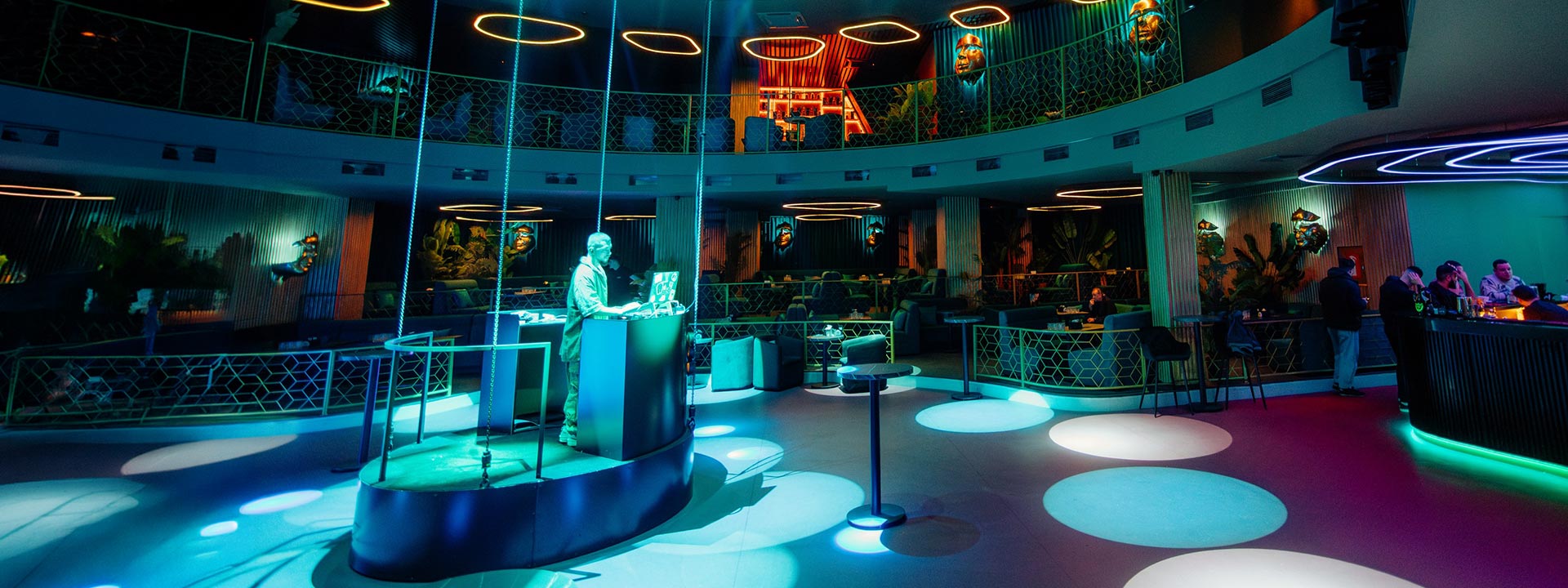 A DJ stands at a booth in the center of a modern, colorful nightclub with circular seating, neon lights, and a few people at the bar.