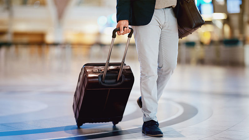 A person in a suit jacket, white pants, and dark shoes walks through an airport with a wheeled suitcase and a shoulder bag.