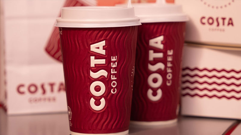Two Costa Coffee cups on a table, with branded packaging in the background
