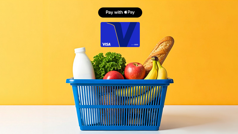 A blue shopping basket with groceries, including milk, lettuce, apples, bananas, and bread, against a yellow background. Above it is a Visa card graphic labeled "Pay with Apple Pay".