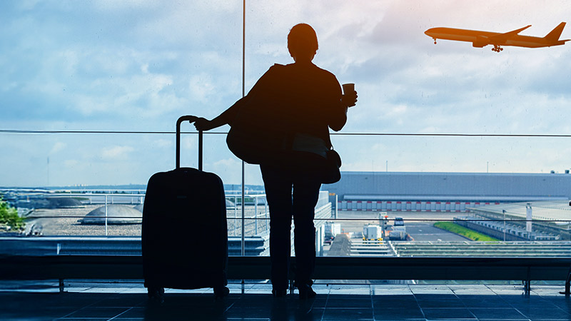 A person stands in an airport terminal with a suitcase, watching an airplane take off through large windows.