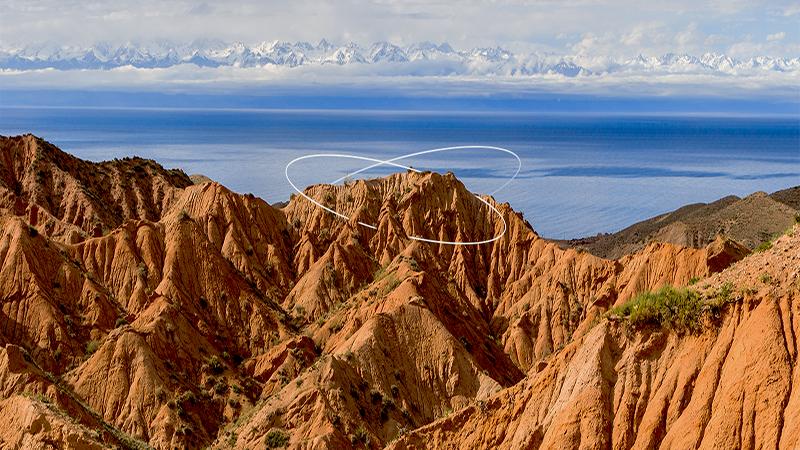 Red rocky hills in the foreground with white lines drawn over a peak, a blue lake in the middle, and snow-capped mountains in the distant background under a cloudy sky.