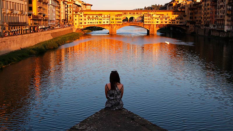 A person sits at the edge of a riverbank overlooking the Ponte Vecchio bridge in Florence, Italy, during sunset.