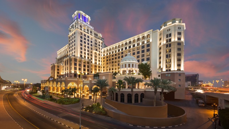 A large, illuminated hotel building at dusk with a dome feature and surrounding palm trees. A road curves alongside, with blurred car lights visible.